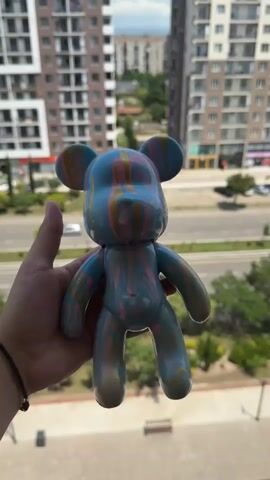A hand holds a toy bear with a marbled pattern of blue, pink, and yellow. The bear is held up in front of a blurred background of apartment buildings and a street with cars.