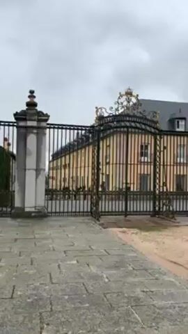 The video shows a large, old, yellow building with white trim behind a tall iron gate.  The sky is overcast.  The camera moves forward and pans to the right, revealing more of the building's facade, which features many windows, decorative trim, and a gray roof with a small tower. The camera then shows a lamppost in the foreground with the building in the background. The area in front of the building appears to be a courtyard paved with sand and some stone.