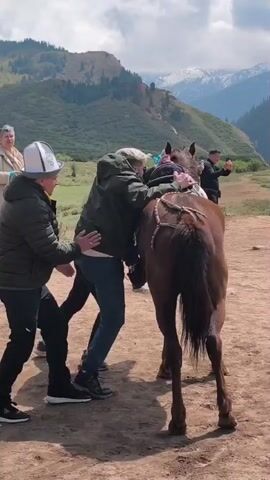 In a wide outdoor shot, several individuals are gathered on a dirt patch, set against a stunning backdrop of green mountains, some of which feature snow-capped peaks, under a cloudy sky. The scene depicts the process of a person mounting a brown horse.

Two men are actively assisting a third person, dressed in a dark green jacket, blue jeans, and dark shoes, in getting onto the horse's back. One man, wearing a black jacket and a distinctive white hat with black patterns, pushes the person's leg up towards the saddle. Another person in a dark vest and cap stands by the horse's flank, also providing support and adjusting the saddle as the rider settles.

After a brief effort, the person successfully mounts the horse, turning to face away from the camera. Other individuals are present in the background: one woman in a light-colored coat stands to the left, observing, and another man further back to the right appears to be filming with a phone. The overall atmosphere seems cooperative and focused on the task of getting the rider securely on the horse in this mountainous region, identified as Jeti-Ögüz, Kyrgyzstan. There is no visible text in the video.