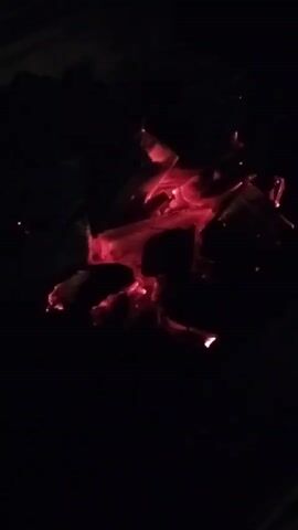 The video shows a close-up, dimly lit view of glowing red coals in darkness. The coals are arranged in a cluster, with some appearing brighter than others. The overall impression is one of embers from a fire.
