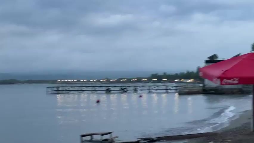 The video shows a tranquil lake scene with a wooden pier extending into the water, illuminated by small lights. A beach area is visible in the foreground, with sand and a few beach umbrellas, one red umbrella with a Coca-Cola logo. The water is calm, with gentle waves lapping at the shore. The sky is overcast, suggesting it might be early morning or late evening. The overall atmosphere is peaceful and serene.