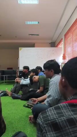 A group of young men are sitting on a green carpeted floor in a room. There is a whiteboard in the background with writing on it. The men are casually dressed and appear to be in a discussion or meeting. One man on the left is holding a phone and speaking. Another man in the center is leaning his head on his hand. The room appears to be a modern office or classroom space with fluorescent lighting and air conditioning units.