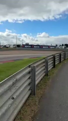 The video shows a car race from a low angle. The camera is behind a grey metal guardrail that runs parallel to the race track. The race track is bordered by green grass with red and yellow painted curbs. A race car, painted in grey, black, and white, speeds by, hugging the inside of the curve. In the background, there is a banner with the word “VAICO” repeated several times. The sky is blue with scattered white clouds. Another race car follows the first one around the bend.