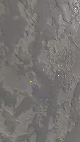 The video captures a close-up view of a textured grey surface that appears to be a wall or a decorative panel. Scattered across the surface are small, iridescent specks that reflect light, creating colorful glints of red, blue, green, and yellow. The texture of the surface itself is uneven, with raised areas and crevices, giving it a rough, almost rocky appearance. The lighting seems to be dynamic, as the glints of color appear and disappear as the camera possibly moves slightly or the light source shifts. There are no people or discernible objects other than the surface itself.