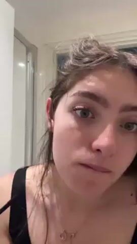 Here is a description of the video:
The video shows a young woman in a bathroom applying makeup to her face. She uses a brush to apply three dots of a dark color under her eye, on her cheekbone. Then, she rubs it in with her finger. The bathroom has a glass shower door. The woman has brown hair and green eyes. She wears a black top and a gold necklace. Her nails are painted with various colors. She is looking directly at the camera and appears serious.