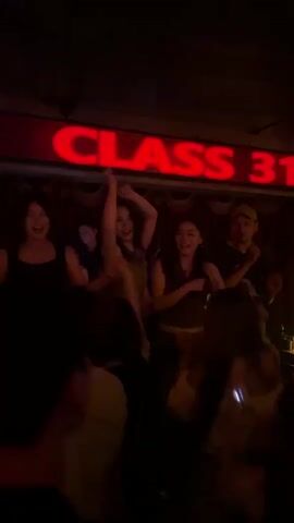 A group of people are dancing on a stage under a neon sign that reads "CLASS 313." The lighting is dim and colorful, with red and purple hues dominating the scene. Several women are visible on the stage, some with their arms raised in the air, appearing to be enjoying themselves. A man wearing a baseball cap is also on stage, seemingly interacting with the dancers. In the foreground, the backs of several other people are visible, suggesting they are part of an audience watching the performance. The overall atmosphere appears to be that of a lively nightclub or party.