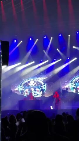 A concert stage bathed in blue and white spotlights hosts a live performance by "Las Hijas De La Cumbia," as displayed on two large screens behind the performers. Red lights are visible near the ceiling. The stage features musical equipment, including a DJ setup. In the foreground, a crowd of spectators enjoys the show, silhouetted against the bright stage. One audience member raises their hand in excitement. Throughout the video, the lighting effects dynamically shift and change.