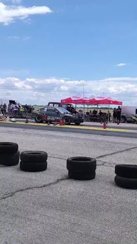 A drag racing car accelerates down a track. The car is black with red and white graphics and appears to be a modified sedan. It is positioned on the left side of the frame, moving from left to right. The track surface is asphalt with white lines. In the foreground, several stacks of used tires are arranged in a line on the edge of the track. In the background, there are tents, people, and other vehicles, suggesting a racing event or pit area. The sky is blue with scattered white clouds, indicating a daytime outdoor setting. The video captures the car's acceleration, with motion blur indicating its high speed.