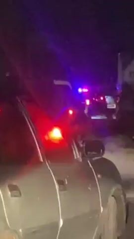 At night, the video shows a gray car with what appears to be tape holding the doors shut. In the background, there are emergency vehicles with flashing red and blue lights. The car is parked on what appears to be the side of the road, and there is a person standing near the emergency vehicles.