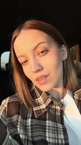 A young woman is applying lip gloss in a car. She has fair skin, green eyes, and shoulder-length blonde hair. She is wearing a white t-shirt and a brown plaid shirt, along with earrings and a necklace. She holds a white tube of lip gloss and applies it to her lips using a small brush applicator. The background is the interior of the car, including the black seat and a glimpse of the outside through the car window. The lighting suggests it is either early morning or late afternoon.