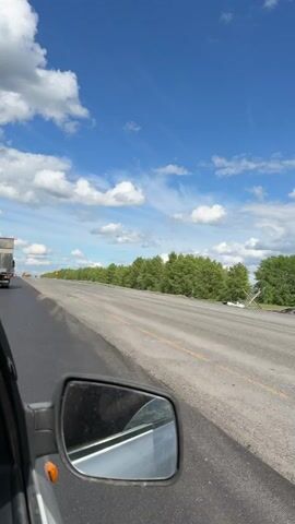 The view from a moving car shows a highway under construction on a sunny day with a blue sky and puffy white clouds. In the left side of the frame is the side view mirror of the car. The highway has fresh, dark pavement on the left lane, while the right lane is older, gray asphalt with yellow lane markings. Red and white striped construction markers are placed along the right side of the road. Beyond the highway is a line of green trees. In the distance, a truck and other vehicles can be seen. The camera appears to be mounted inside the car, capturing the roadside view during the drive.