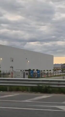 The video shows a white building with a metal fence in front of it. There are trees and grass between the fence and the road. The building has several lights on it and what appears to be a logo on the right side. The sky is cloudy. The video is taken from a moving vehicle.