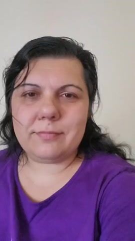 This is a short, close-up video of a woman with dark, curly hair and fair skin. She is wearing a purple t-shirt. The background is a plain, light-colored wall. The woman is looking directly at the camera and occasionally smiles. The camera is held at eye level and is relatively still, though there are some slight movements and blurs. The lighting appears to be natural and soft.