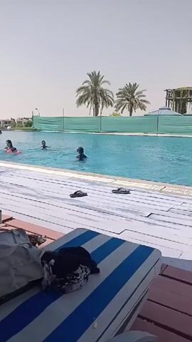 The video shows a swimming pool with several people swimming. There are lounge chairs in the foreground with striped cushions. The weather appears to be sunny.