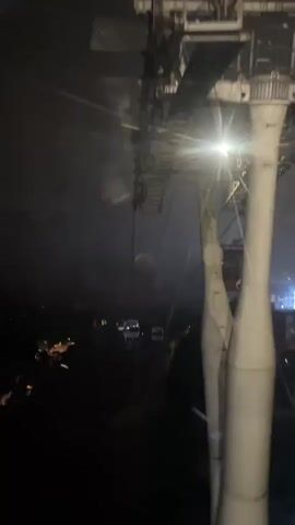 The video shows a view from inside a cable car at night. The cable car is moving, as other cable cars are seen further down the line. The scene is dark, with the ground below barely visible. Some lights from buildings and vehicles can be seen. The structure of the cable car system is visible above, with wires and supports illuminated by a bright light. The overall atmosphere is foggy and slightly blurry. Voices can be heard in the background.