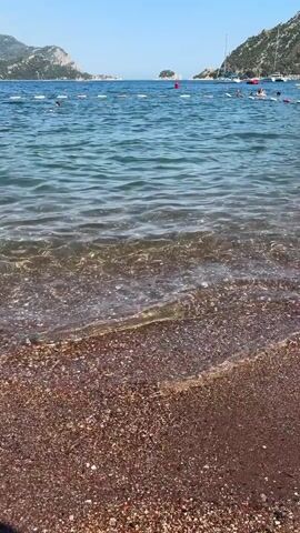 The video opens with a view of a beach and the ocean. The camera is angled downwards towards the shore, where small waves are gently lapping at the pebbles. The water is clear and blue, with sunlight glinting off the surface. In the background, a calm sea stretches out towards a coastline dotted with green hills and mountains. Several buoys mark a swimming area, and in the distance, a few boats and people can be seen in the water. The sky is clear and bright blue, indicating a sunny day. The scene appears tranquil and idyllic, suggesting a peaceful day at the beach.
