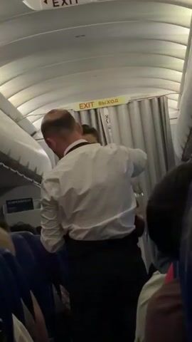 A medium shot shows the interior of an airplane cabin from the perspective of a passenger. The focus is on two people in the aisle, appearing to be an argument or altercation. A man in a white dress shirt and dark pants, possibly a flight attendant, is facing away from the camera and appears to be restraining or interacting with a woman in a red garment. Her face is partially obscured. They are positioned near an exit door, indicated by illuminated signs with the word "EXIT" and its Russian translation, "ВЫХОД". The aisle is lined with seats, some occupied by other passengers whose backs are to the camera. The overall lighting is dim, typical of an airplane cabin, with overhead lights contributing to the illumination.
