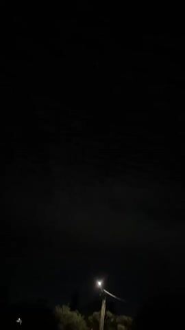 The video shows a dark night sky with some faint outlines of clouds. There is a single street light visible in the lower right corner of the frame, illuminating a small area of the sky. The rest of the scene is completely dark.