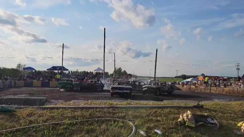 A demolition derby event is taking place on a dirt track. Several cars, in various states of disrepair, are driving around the track, some colliding with each other. There is a crowd of spectators gathered around the track, watching the event. The sky is blue with some clouds, and the sun is shining. In the foreground, there is a grassy area with some hoses and equipment scattered on the ground.