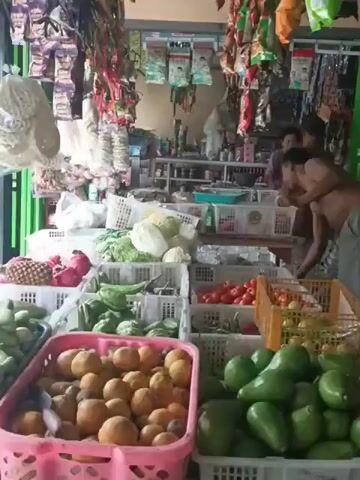 THE VIDEO SHOWS A MAN, BARE-CHESTED AND WEARING SHORTS, STANDING IN WHAT APPEARS TO BE A SMALL GROCERY STORE OR MARKET STALL. HE IS HOLDING A PLASTIC BAG AND APPEARS TO BE ENGAGED IN STOCKING OR ARRANGING ITEMS. IN THE BACKGROUND, ANOTHER PERSON, WEARING A BROWN SHIRT, IS VISIBLE BEHIND A COUNTER FILLED WITH VARIOUS PRODUCTS. THE STALL IS STOCKED WITH A VARIETY OF FRUITS AND VEGETABLES DISPLAYED IN BASKETS, INCLUDING AVOCADOS, ORANGES, TOMATOES, AND CABBAGE. SHELVES BEHIND THE COUNTER ARE FILLED WITH PACKAGED GOODS AND BOTTLES. THE ENVIRONMENT SUGGESTS A COMMERCIAL SETTING, LIKELY OUTDOORS OR IN A SEMI-OPEN-AIR STRUCTURE DUE TO THE NATURAL LIGHTING AND THE PRESENCE OF HANGING MERCHANDISE. THE OVERALL ATMOSPHERE IS THAT OF A TYPICAL MARKET SCENE.