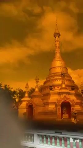 The video showcases a golden pagoda with an ornate design, set against a striking orange sky. The camera then pans across a green lawn to reveal a traditional Thai building with a red roof. A man with a mustache, wearing a camo bandana and a crossbody bag, appears in front of the building. The sky retains its orange hue throughout the video.