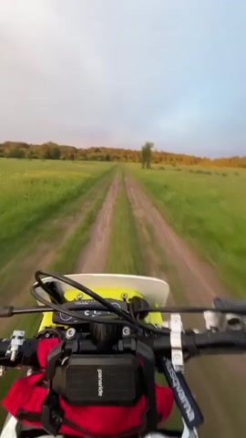 A first-person point-of-view video shows someone riding a yellow-and-white dirt bike on a dirt road through a grassy field. The road has two tracks and leads towards a line of trees in the distance. The sky is overcast. The handlebars of the bike are visible. A small red bag is attached to the bike.