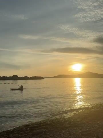 The video shows a sunset over a body of water, possibly a sea or lake. In the foreground, there is a pebble beach with gentle waves lapping at the shore. The sun is low on the horizon, casting a warm golden glow across the water and sky. Wispy clouds are scattered across the sky. In the middle ground, a person is kayaking on the water. A line of buoys is visible, marking off an area of the water. In the background, there are hills or mountains. The overall mood of the video is peaceful and serene.