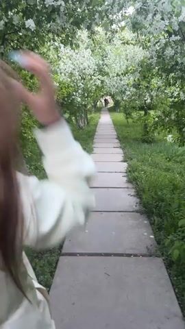 A woman walks along a concrete path lined with blooming trees. She is wearing a light-colored tracksuit, sneakers, and sunglasses. The path is straight and leads towards a distant figure at the end. The trees on either side of the path are full of white flowers. Green grass and plants grow on the ground around the path. The woman turns and smiles at the camera, waving.