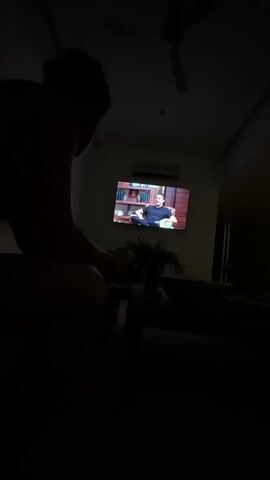 A person sits in a dark room, watching a TV show. The person is silhouetted, and their features are not clearly visible, though it appears they are male and have dark hair. The TV screen displays a man sitting in a chair in front of a bookcase, and he is speaking. There is a plant on a table in the room, and a ceiling fan is visible above. The scene is dimly lit.