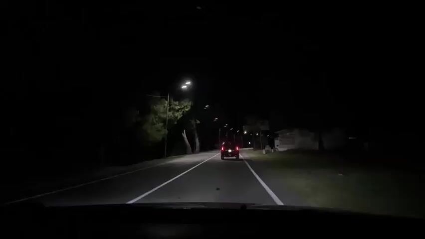 A first-person perspective video shows a car driving on a dark, paved road at night. The car's headlights illuminate the road ahead, revealing two white lines dividing the lanes. In the distance, another car with red taillights is visible, also traveling in the same direction. Streetlights line the right side of the road, casting pools of light on the asphalt and the surrounding vegetation. Trees with sparse foliage are visible on the left side of the road. On the right side of the road, there is a small, dilapidated building with a corrugated metal roof and a window. The dashboard of the car is visible in the foreground, displaying the time as 8:10 and the temperature as 67 degrees Fahrenheit. The overall scene depicts a nighttime drive through a sparsely populated area with some signs of human habitation.