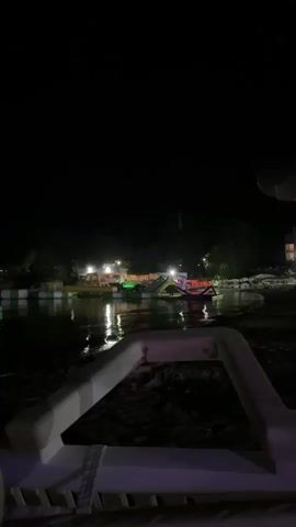 The video opens with a close-up of a white object in the foreground, which appears to be a lounge chair. The camera then pans out to reveal a body of water at night, with several inflatable structures floating on it. Lights are visible on the shore, illuminating the structures and the water's surface. The overall atmosphere is dark and somewhat festive, with the lights creating reflections on the water. There are no people clearly visible in the frame, but the presence of the inflatable structures suggests a recreational area that is open at night. The environment appears to be outdoors, likely a park or a resort with a water feature.