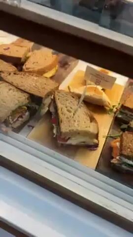 The video shows the interior of a cafe display case. The top shelf displays sandwiches on wooden boards, with small signs identifying the contents. Below, there are individual plastic cups containing fruit and granola. There are also cans of cold brew coffee. The bottom shelf showcases various cakes and pastries, including layered chocolate cake, several muffins or small cakes with toppings, brownies, and a tart or pie. The shelves are well-lit and clean. The overall setting is a bright and inviting cafe environment. The camera moves slowly, providing a view of the various food items available.