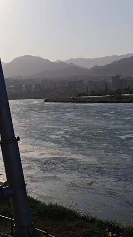 A wide, choppy river flows in the foreground. The water is a pale blue-green with white foamy patches. In the background, a city skyline rises behind a tree-lined embankment. Buildings vary in height and architectural style, with some appearing to be under construction. Mountains form a hazy backdrop to the city, their peaks softened by distance and atmosphere. Several boats are docked along the embankment. Two small speedboats are moored alongside a larger, white-and-blue yacht. A cable car system is visible in the distance, with gondolas suspended in the air. The sky is a pale, washed-out blue, indicating a bright but perhaps hazy day.