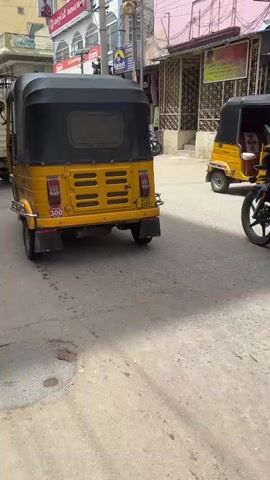 A yellow auto-rickshaw with a black canopy is parked on the left side of a street. A man wearing a white shirt and pants is riding a black motorcycle away from the camera. The motorcycle has a yellow license plate. On the right side of the street, there are shops with signs in Telugu. One sign says "VASAVI NIVAS - PUTTAPARTHI". Another sign advertises "SAMSUNG". Several motorcycles are parked in front of the shops. The street is paved with asphalt and has some cracks and patches. The buildings on the right are multi-story with colorful facades. The sky is overcast.