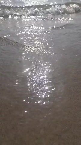 A very close-up shot of a gentle wave washing onto a sandy beach. The sun glints off the water's surface, creating shimmering patterns on the wet sand. The water is clear and shallow, and the sand appears to be finely grained. No people or objects are visible in the frame. The overall mood is peaceful and serene.