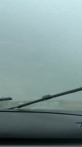 The video shows a view from inside a car driving on a highway during a rainstorm. The windshield is covered in raindrops, distorting the view of the road ahead. Several other vehicles, including trucks, can be seen on the highway, and an overpass is visible in the distance. The sky is overcast and gray.