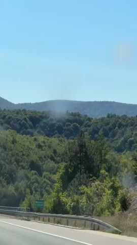 This video shows a view from a vehicle driving on a highway. The highway has a metal guardrail on the right side, and it is bordered by lush green trees and hills. In the distance, there are mountains under a clear blue sky. There is a sign on the side of the road with green text, but it is not clearly legible. A small white vehicle can be seen driving ahead on the highway. The video appears to be filmed during the daytime.