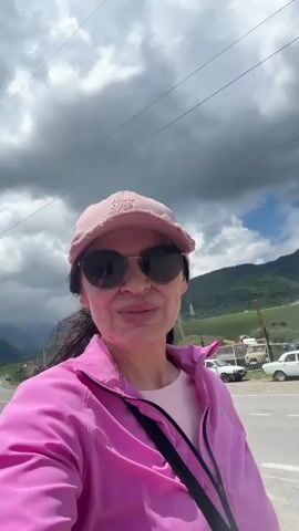 A woman is filming herself in a selfie video. She is wearing a pink baseball cap, sunglasses, and a bright pink jacket over a pink shirt. She is standing on the side of a paved road, with mountains in the background. The sky is cloudy. She smiles and gestures with her right hand. Cars and some buildings are visible in the background.
