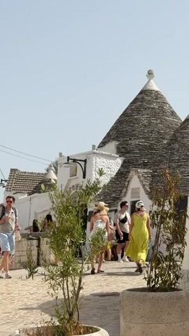 The video shows a street scene with several people walking around in what appears to be a historic town. The architecture is unique, with conical-shaped roofs that are characteristic of the trulli houses found in Puglia, Italy. The buildings are made of white stone and the roofs are covered in gray stone tiles. Some of the buildings have small shops or cafes. The street is paved with cobblestones. The people are dressed in casual summer clothing. Some of the people are carrying backpacks, suggesting they are tourists. The sky is clear and the sun is shining, indicating it is a warm day. There are some plants and trees scattered throughout the scene, adding greenery to the environment. One of the buildings has a display cabinet mounted on the wall, showcasing items for sale. Another building has a ladder leaning against it, possibly for maintenance. The overall impression is of a lively tourist destination with a distinctive architectural style.