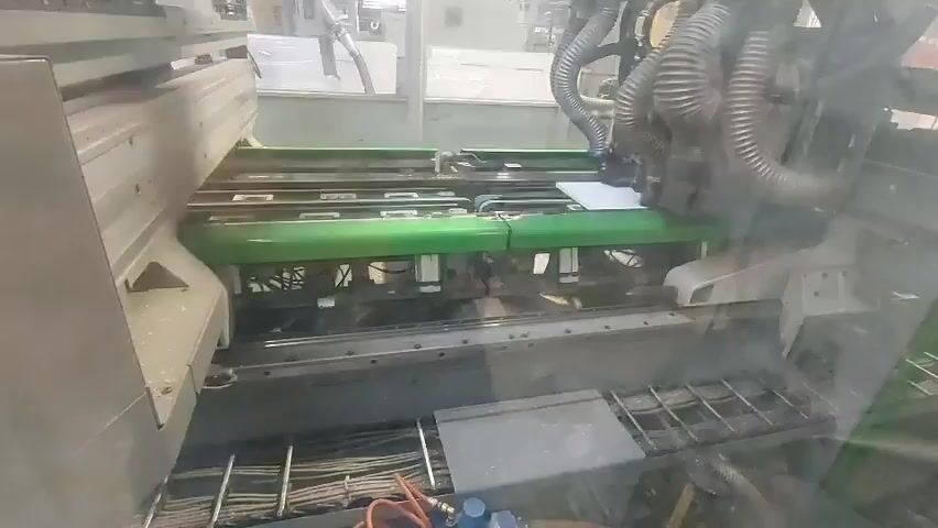 The video shows a close-up view of the interior of an industrial machine, possibly a printer or binder. A white sheet of paper is visible being fed through the machine on a conveyor belt. The machine has several green components and a complex system of tubes and wires. The overall environment appears to be a factory or workshop setting, with other machinery visible in the background through a clear protective barrier. The lighting is adequate to see the details of the machine's operation.