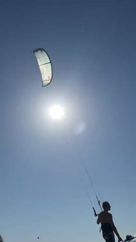 In this media file, a kitesurfer is visible against a clear blue sky, with the sun shining brightly overhead. The kitesurfer, a silhouette against the sun, is holding onto a control bar connected to a large, green and white kite. The kite is positioned high in the sky, catching the wind. The sun is positioned centrally in the frame, creating a lens flare on the right side. In the lower part of the frame, a tiny bird is seen flying. The scene suggests a sunny day, likely during daylight hours, with the activity of kitesurfing taking place. The location is identified as Petten, Netherlands. The overall impression is one of outdoor recreation and activity under a bright sky.
