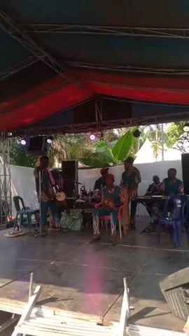 A music band of seven members plays music on a stage. They are all wearing teal and orange patterned outfits. The stage has a metal frame overhead, covered with red and dark blue material. There is a drum set, a keyboard, and several men playing various hand percussion instruments. Behind the stage is a white panel, and trees are visible in the background. The stage is made of gray tiles.