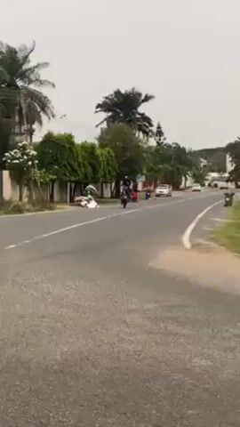 The video shows a man riding a motorcycle on a paved road lined with trees and other vegetation on both sides. The motorcycle is moving towards the camera and away from a group of people standing by the side of the road. There are a few cars parked further down the road. The sky is overcast and the general environment suggests a tropical or subtropical location.