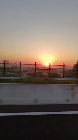 The video shows a sunset viewed from a moving vehicle, likely on a highway. The sun is partially obscured by trees and a fence, casting an orange glow across the sky. The foreground shows the road and a concrete barrier. The camera is angled to capture the horizon and the sky. The video is short, lasting only a few seconds, and appears to be a continuous shot.