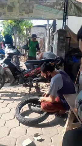 The video shows a street scene with several people. In the foreground, a person wearing a blue shirt and red pants is kneeling and working on a motorcycle tire. Next to them is another tire. Behind them, a person in a gray shirt is standing and observing. To the left, a motorcycle is parked. In the background, two more individuals are visible, one in a green shirt and another in a blue shirt and dark vest. The environment appears to be an outdoor workshop or roadside repair area, with a paved ground and some structures in the background. The overall lighting suggests it is daytime.