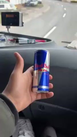 The video shows the interior and the point of view from inside a vehicle, likely a truck, as it drives through a residential street. Initially, a hand holds a can of Red Bull energy drink. The camera angle shifts to show the road ahead, lined with houses. The vehicle continues driving slowly through the neighborhood. Then, the hand places the Red Bull can on the dashboard, and in its place, a green can of Fanta drink appears.