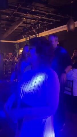 A woman in a white pleated dress with a black belt is dancing. She has dark hair and is smiling. She is in a dimly lit room with red and blue lights. There are other people in the background, some of whom are also dancing. The environment appears to be a party or a club.