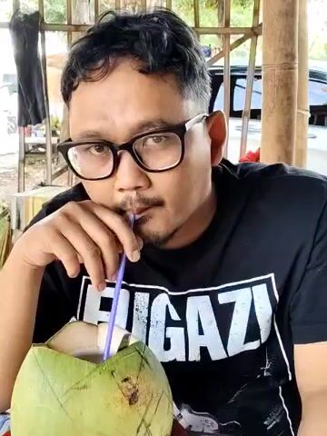 A man in a black "FUGAZI" t-shirt and glasses is drinking from a green coconut with a blue straw. He is sitting under a partially covered outdoor structure made of bamboo. The environment appears to be somewhat rural or semi-rural, with trees and a vehicle visible in the background. The man looks directly at the camera at several points during the video.