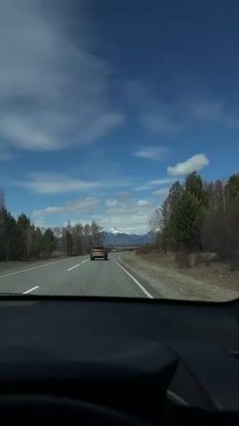 The video captures a first-person perspective from inside a moving car, looking out through the windshield. The car is traveling on a two-lane paved road with white lane markings. Ahead, another car, light yellow or tan in color, is visible in the distance. The sides of the road are lined with a mix of deciduous trees, some bare and some with light green foliage, and evergreen trees. Beyond the trees, in the far distance, snow-capped mountains rise under a bright blue sky dotted with scattered white clouds. The overall scene suggests a clear, sunny day in a rural or mountainous region. No people are directly visible, and there is no discernible text. The setting is Nikol'sk, Russia.