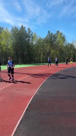 The video shows several individuals running on an outdoor track. The track is composed of red running lanes and a black perimeter. A group of trees lines the background of the track, and the sky above is blue with scattered clouds. Runners are wearing athletic attire and are spaced apart as they proceed along the track.